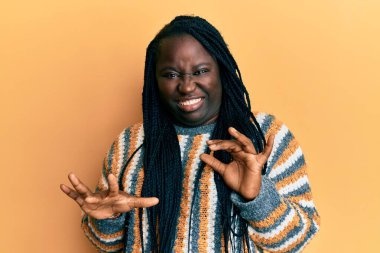 Saçları örgülü genç siyahi bir kadın. Kış süveteri giymiş. Tiksindirici tepki verdiği için rahatsız ve korkmuş bir ifade takınıyor.. 