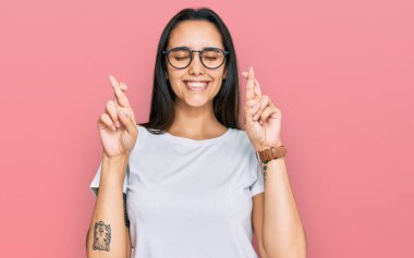 Latin kökenli genç bir kadın, günlük beyaz tişört giyip umut ve gözlerin kapalı bir şekilde parmaklarını çaprazlayarak işaret ediyor. Şans ve batıl inançlar. 