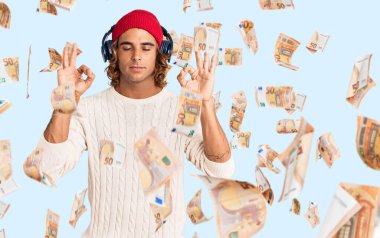 Kulaklık kullanarak müzik dinleyen, gözleri kapalı gülümseyen genç İspanyol adam parmaklarıyla meditasyon hareketi yapıyor. yoga konsepti.