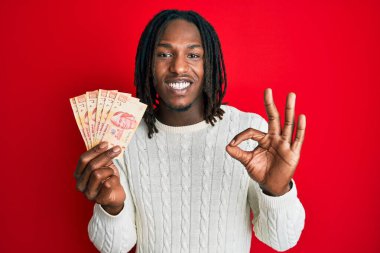 Saçları örgülü Afrikalı Amerikalı bir adam 100 Meksika pezosu tutuyor. Parmaklarıyla imza atıyor, gülümsüyor ve mükemmel bir sembol. 