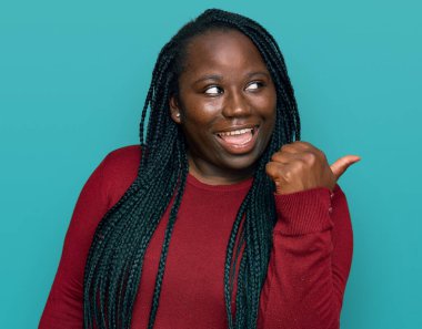 Örgülü, günlük kıyafetler giyen, mutlu yüzlü, gülümseyen ve baş parmağıyla yüzünü gösteren genç siyahi bir kadın.. 