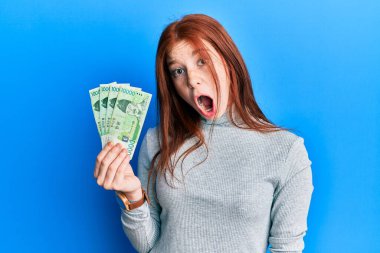 Young red head girl holding 10000 south korean won banknotes scared and amazed with open mouth for surprise, disbelief face 