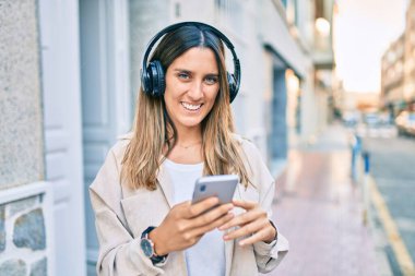 Beyaz kadın şehirde akıllı telefon ve kulaklık kullanarak mutlu bir şekilde gülümsüyor..