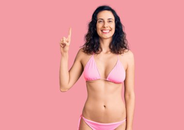 Genç ve güzel İspanyol kadın bikini giyiyor ve bir numaralı parmağını gösterip işaret ederken kendinden emin ve mutlu gülümsüyor.. 