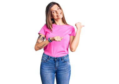 Güzel, beyaz bir kadın, günlük kıyafetler giyip arka tarafı işaret ediyor. Elleri havada, kendinden emin gülümsüyor. 