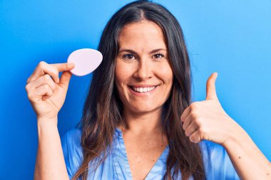 Genç, güzel esmer kadın makyaj yapıyor mavi arka planda sünger kullanıyor mutlu ve pozitif gülümsüyor, başparmağını kaldırıp mükemmel ve onay işareti yapıyor.