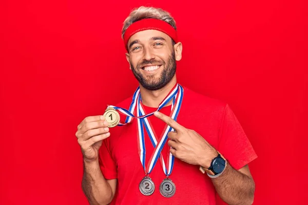 若いですハンサムなブロンドスポーツマンとともにあごひげ受賞メダル上の隔離された赤背景笑顔幸せなポインティングとともに手と指 — ストック写真