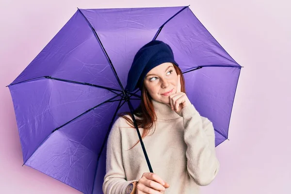 若いです赤頭の女の子保持紫色の傘身に着けています新鮮なベレー帽深刻な顔考えについて質問に手であご 気になるアイデア — ストック写真