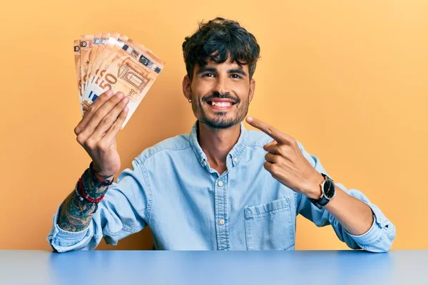Joven Hispano Sosteniendo Billetes Euros Sentado Mesa Sonriendo Feliz Señalando —  Fotos de Stock
