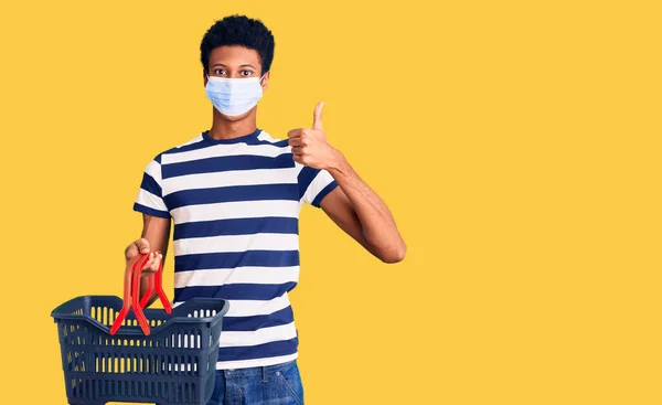 若いアフリカ系アメリカ人の男を着てショッピングバスケットと医療マスク笑顔と正 親指アップ行う優れた承認サイン — ストック写真