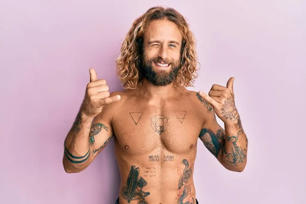 Handsome man with beard and long hair standing shirtless showing tattoos doing shaka sign winking looking at the camera with sexy expression, cheerful and happy face. — Stock Image Handsome Man Beard Long Hair Standing Shirtless Showing Tattoos Doing Stock Picture