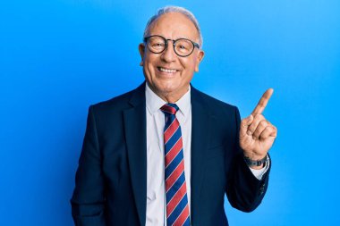 İş elbisesi giyen ve yüzünde kocaman bir gülümseme olan beyaz bir adam, kameraya el ile işaret ediyor.. 