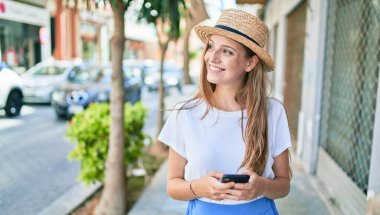 Genç sarışın kadın tatilde gülümsüyor. Şehrin caddesinde akıllı telefon kullanıyor.