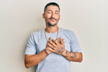 90 'lı yılların tarzında dövmeleri olan, elleri göğsünde gülümseyen, yüzünde minnet ifadesi olan yakışıklı bir adam. sağlık kavramı. 