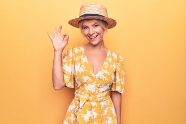 Yaz şapkası takıp sarı arka planda giyinen güzel sarışın kadın el ve parmaklarıyla tamam işareti yaparken gülümsüyor. Başarılı ifade.