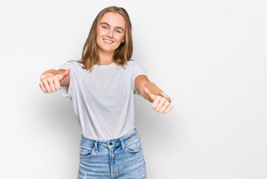 Güzel, genç, sarışın bir kadın, günlük beyaz tişört giyip olumlu bir jest yapmayı onaylıyor, gülümsüyor ve başarı için seviniyor. kazanan jest. 