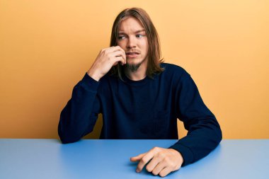 Uzun saçlı, sıradan giysiler giyen yakışıklı beyaz adam masada oturmuş gergin ve elleri ağızdan tırnak yerken gergin görünüyor. anksiyete. 