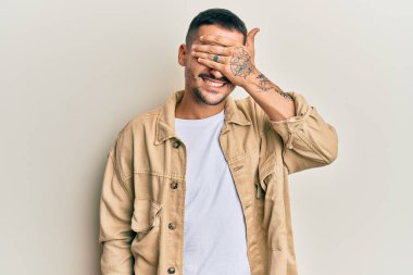 Dövmeleri olan, sıradan kahverengi kot ceket giyen, gülümseyen ve elleri yüzünde, şaşırtmak için gözlerini kapatan yakışıklı bir adam. kör kavram. 