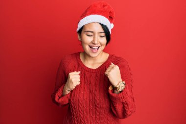 Kısa saçlı güzel bir kadın Noel şapkası takıyor. Şaşırmış ve şaşırmış bir şekilde kolları havada ve gözleri kapalı bir şekilde başarıyı kutluyor. 