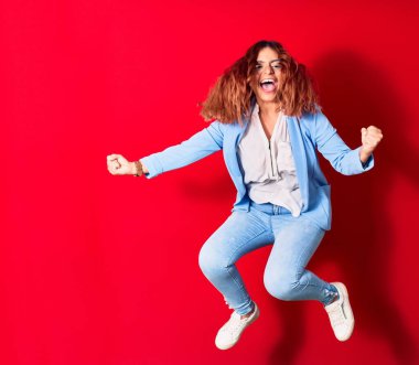 Genç, güzel, Latin bir kadın günlük kıyafetler giyip mutlu gülümsüyor. Yüzünde tebessümle zıplıyor, kırmızı arka planda yumruklarıyla kazanan jestler yapıyor.