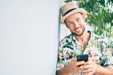 Beyaz tenli yakışıklı adam dışarıda mutlu bir şekilde gülümsüyor. Duvara yaslanmış akıllı telefon kullanıyor.