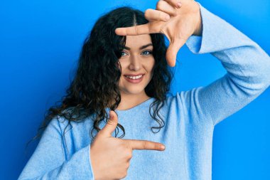 Genç esmer, kıvırcık saçlı, günlük kıyafetler giyen, gülümseyen, elleri ve parmakları mutlu yüzlü bir kadın. yaratıcılık ve fotoğrafçılık kavramı. 