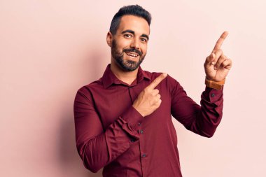 Gündelik kıyafetler giyen genç İspanyol adam gülümseyip kameraya bakıyor ve iki eliyle ve parmaklarıyla kenarı işaret ediyor.. 