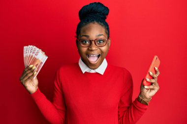 Genç Afro-Amerikalı kadın elinde akıllı telefon ile Norveçli Krone banknotları tutuyor. Çılgınlığı kutluyor ve başarıyı hayretle kutluyor.. 