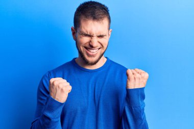 Gündelik kazak giyen genç yakışıklı adam şaşkınlık ve şaşkınlık içinde kollarını kaldırarak ve gözlerini kapayarak başarıyı kutluyor. 