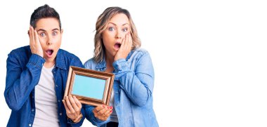 Couple of women holding empty frame afraid and shocked, surprise and amazed expression with hands on face 