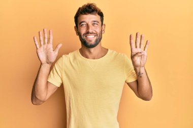 Sarı arka planda sarı tişört giyen sakallı yakışıklı bir adam dokuz numaralı parmaklarını gösterip gülümserken mutlu ve kendinden emin.. 