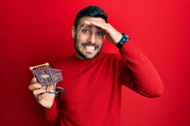 Elinde küçük bir süpermarket arabası tutan İspanyol bir genç. Kafası karışık, şaşırmış ve sinirli bir yüzü var. 