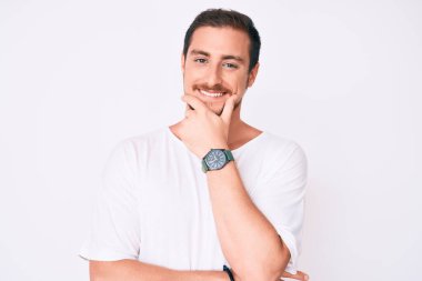 Gündelik beyaz tişört giyen genç yakışıklı kameraya kendinden emin bir şekilde bakıyor kollarını kavuşturmuş ve çenesini kaldırmış gülümsüyor. Olumlu düşünmek. 