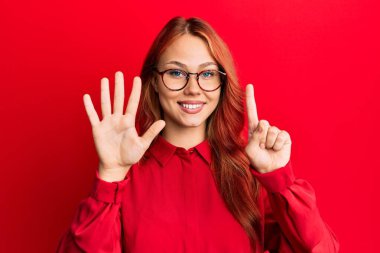 Kırmızı arka planda günlük kıyafetler ve gözlük takan genç, güzel bir kızıl kadın. Altıncı parmaklarıyla işaret ederken kendinden emin ve mutlu bir şekilde gülümsüyor.. 