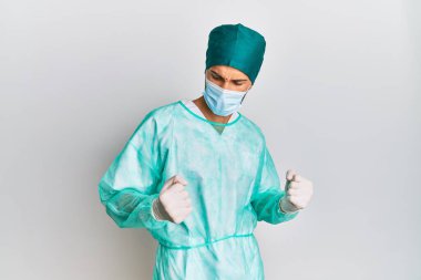 Cerrah üniforması ve tıbbi maske takan yakışıklı genç adam çok mutlu ve heyecanlı. Kolları havada, gülümseyen ve başarı için bağıran kazanan bir jest yapıyor. kutlama konsepti. 