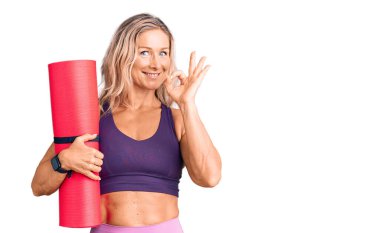 Orta yaşlı, zinde, yoga minderi tutan sarışın kadın parmaklarıyla imza atıyor, gülümseyerek mükemmel bir sembol oluşturuyor. 