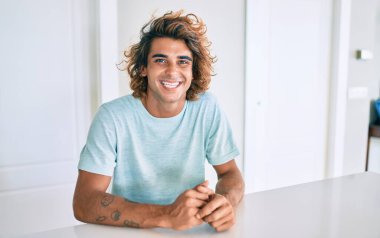 Genç İspanyol adam mutlu bir şekilde gülümsüyor. Evdeki masada oturuyor.