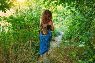 Orta yaşlı İspanyol kadın, sıradan kıyafetler ve sırt çantası giyerek yeşil bitkilerin etrafındaki kırsal yolda yürüyor.