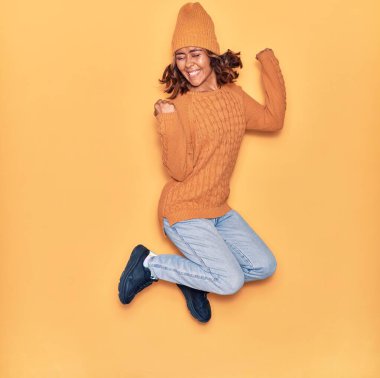 Yün şapka ve süveter giyen, mutlu gülümseyen genç, güzel Latin bir kadın. Yüzünde tebessümle zıplıyor, sarı arka planda yumruklar havada kutluyor.