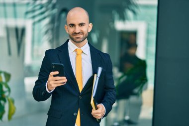 Genç Latin kökenli kel iş adamı şehirde akıllı telefon kullanarak mutlu bir şekilde gülümsüyor..