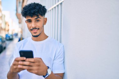 Genç Arap adam mutlu bir şekilde gülümsüyor. Duvara yaslanmış akıllı telefon kullanıyor..