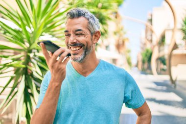 Orta yaşlı, kır saçlı, akıllı telefondan konuşan bir adam şehrin caddesinde yürüyor..