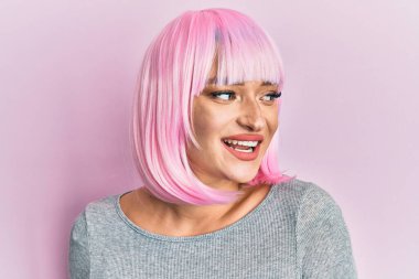 Pembe peruklu genç beyaz kadın yüzünde gülümsemeyle yan yana bakıyor, doğal bir ifade. Kendinden emin gülen. 