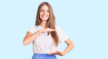 Yaz tarzı giyinen genç, sarışın bir kadın. Elleriyle büyük ve büyük bir işaret gösteriyor. Ölçü sembolü. Kameraya bakıp gülümsüyor. ölçüm kavramı. 