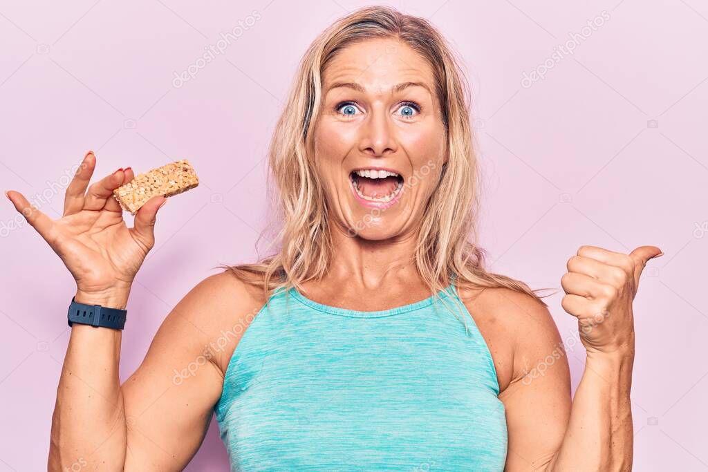 Middle age caucasian blonde woman eating protein bar as healthy energy snack pointing thumb up to the side smiling happy with open mouth 