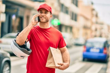 Beyaz atlı genç teslimatçı akıllı telefondan konuşuyor ve şehirde kağıt torba taşıyor..