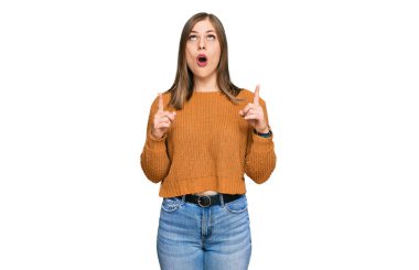 Gündelik giysiler giyen güzel beyaz kadın şaşkın şaşkın yukarı bakıyor ve parmaklarıyla işaret ediyor ve kollarını kaldırıyor.. 