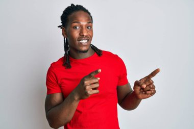 Saçları günlük kıyafetlerle örülmüş, gülümseyen ve kameraya bakıp iki eliyle ve parmaklarıyla kenarı gösteren Afro-Amerikan bir adam.. 