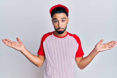 Young man with beard wearing baseball uniform clueless and confused with open arms, no idea and doubtful face. 