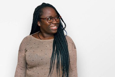 Saçları örgülü ve gözlüklü genç siyahi bir kadın yüzünde gülümseme, doğal bir ifade ile yan yana bakıyor. Kendinden emin gülen. 